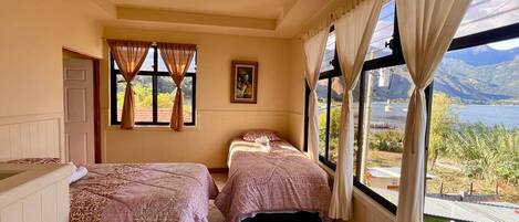 A bedroom with two beds, a window with curtains, a painting on the wall, and a view of mountains and water.