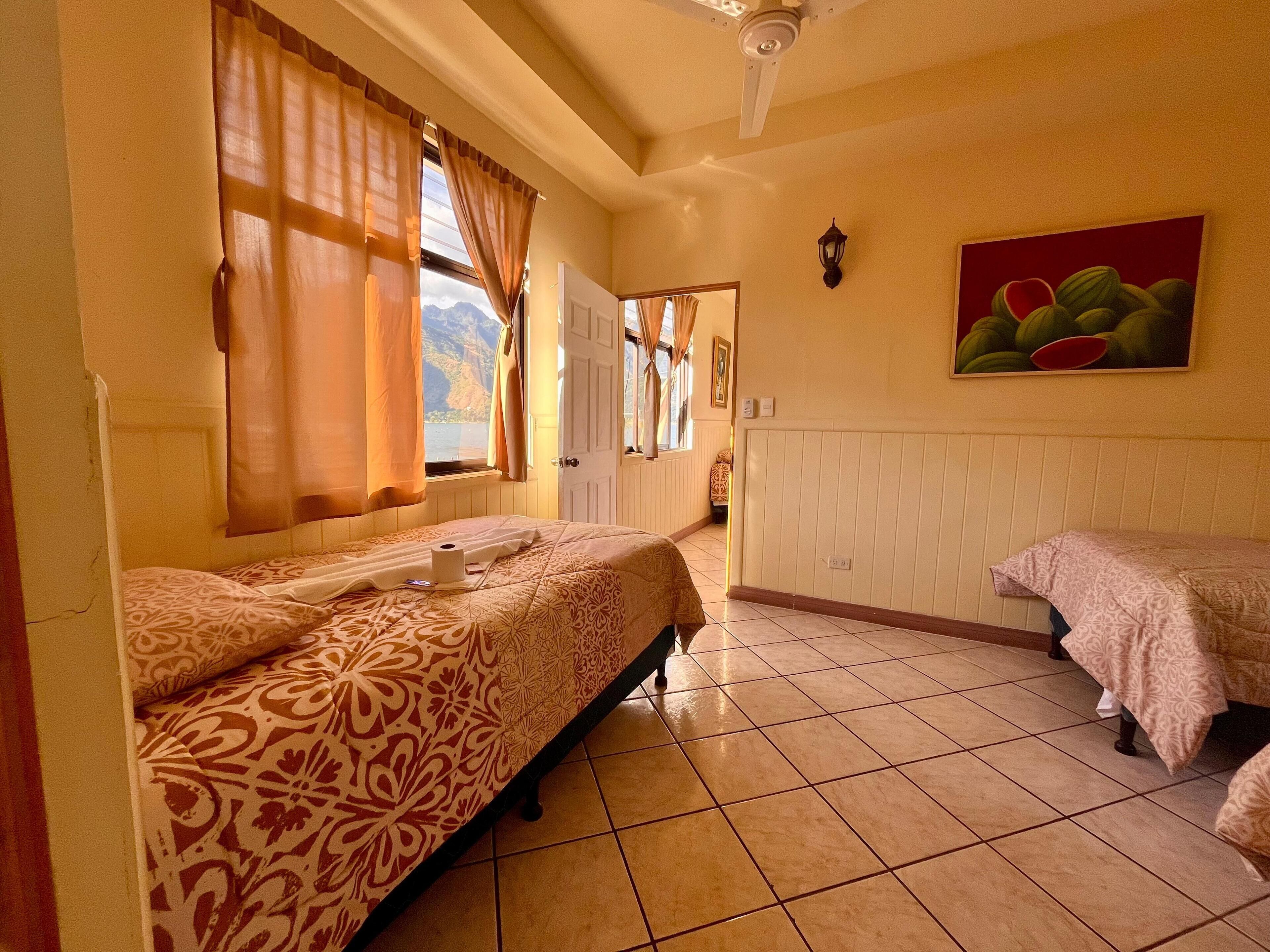 A hotel room with two beds, a view of the mountains through the window, a painting on the wall, and a ceiling fan.