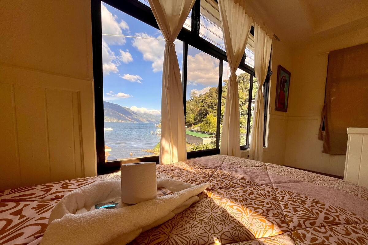 A bedroom with a large window offering a view of a lake and mountains.