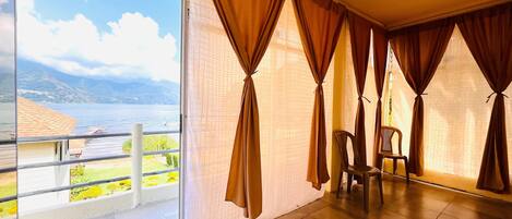 A room with a large sliding glass door leading to a balcony with a view of a lake and mountains. There is a chair and a bed in the room.