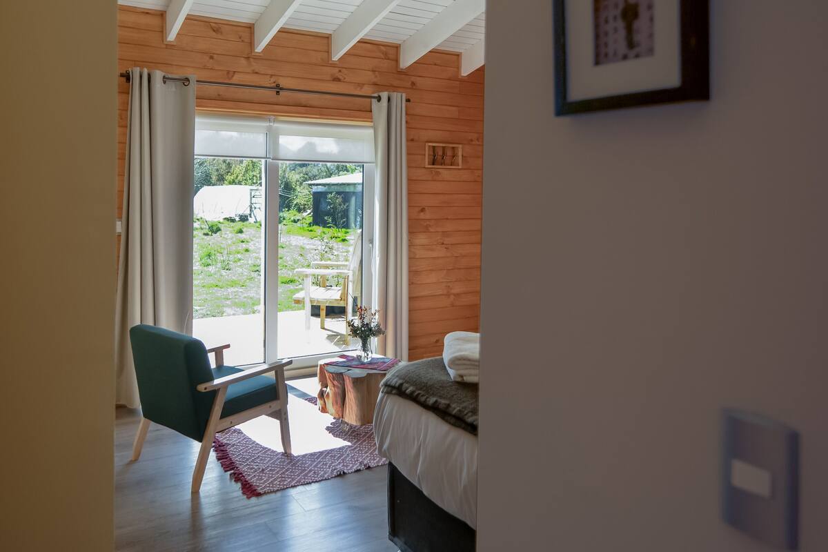 Un dormitorio con cama, una silla, una mesita y vistas al exterior por un ventanal corredizo.