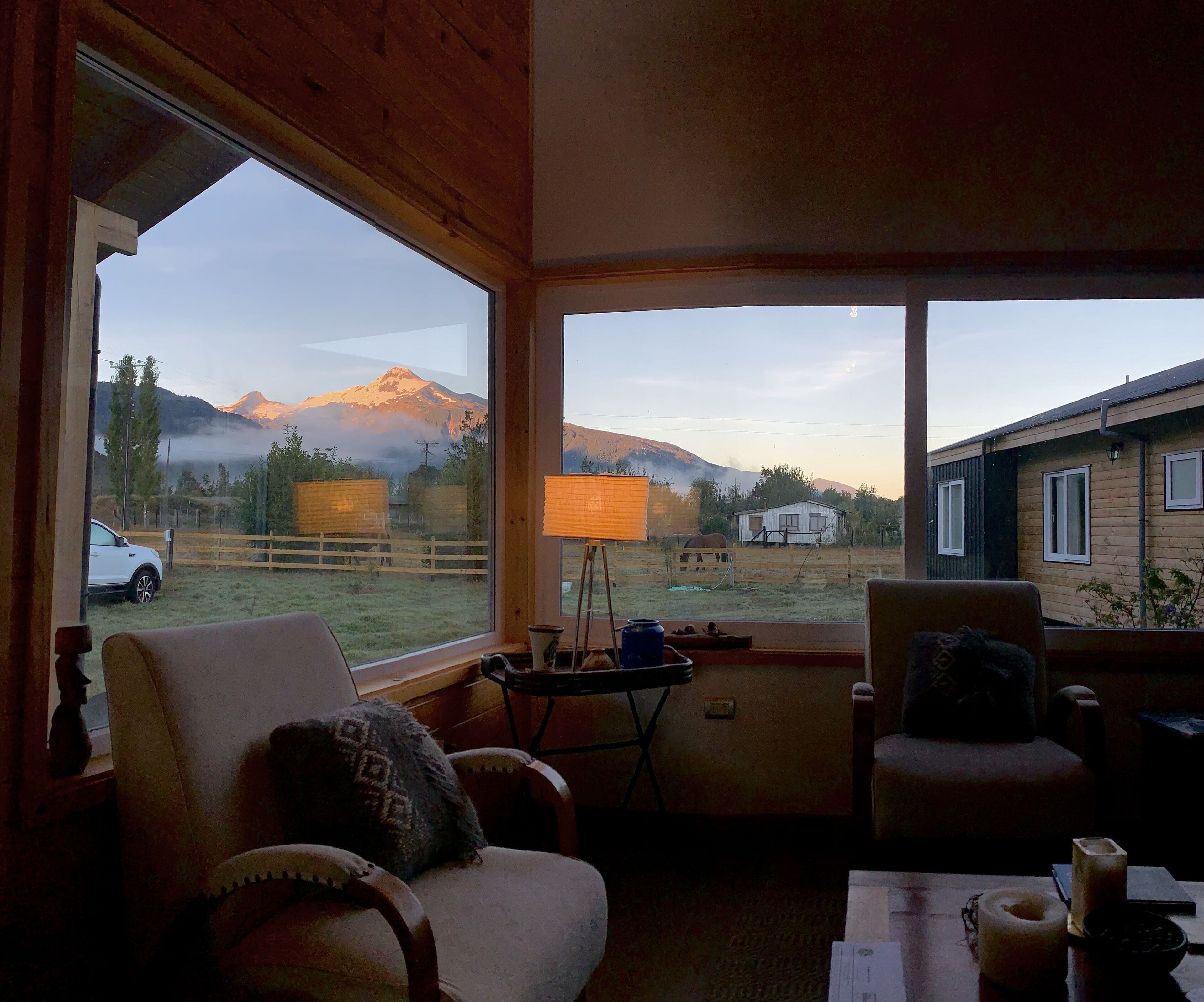 Une pièce chaleureuse avec une grande fenêtre donnant sur une montagne et un chalet en bois. On y trouve deux fauteuils et une petite table avec une lampe.