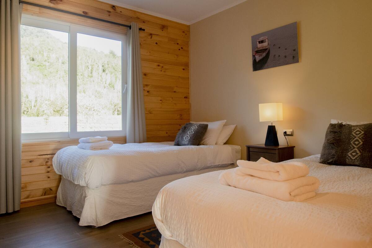 Una habitación con dos camas, una ventana con cortinas, un cuadro en la pared y una lámpara de noche.