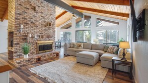 Un salon spacieux avec une cheminée en briques, un plancher en bois, un grand canapé d’angle et une vue agréable par les fenêtres.