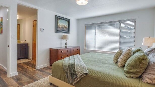 Une chambre à coucher comprenant un lit, une commode, une fenêtre avec des stores, un tableau accroché au mur et une porte donnant sur une autre pièce.