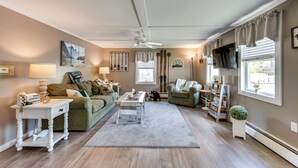 A living room with a sofa, a coffee table, a TV, and a ceiling fan. - Steps to Crystal Lake: Spacious Retreat w/ Views (Barton)