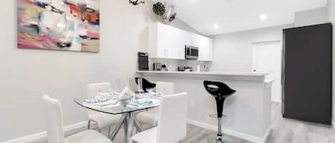 Une cuisine moderne avec des meubles blancs, un îlot central et un coin repas comprenant une table en verre et des chaises blanches.