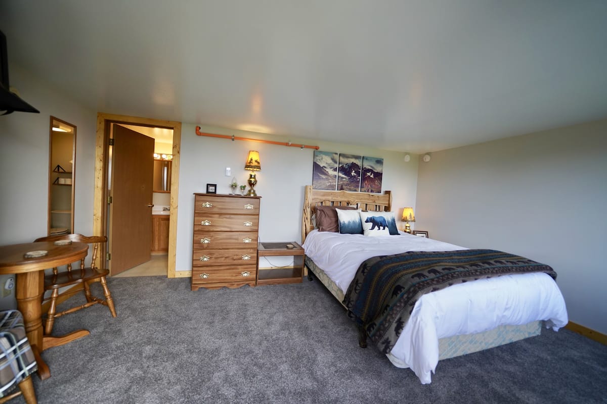 A bedroom with a bed, dresser, nightstand, lamp, and a painting on the wall.