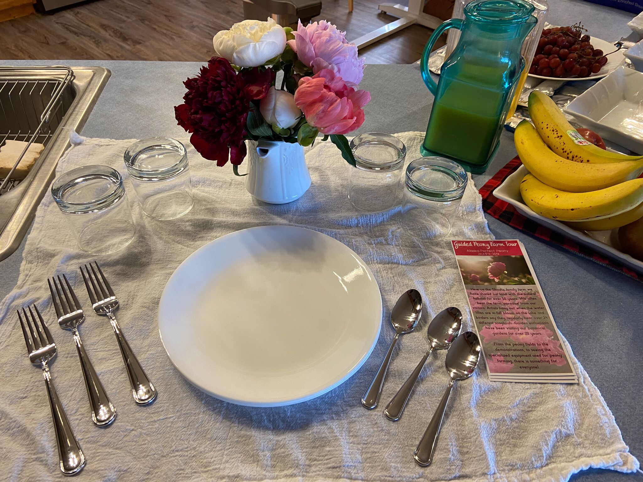 A table set with a white plate, silverware, a vase with flowers, a pitcher of green juice, and a brochure.