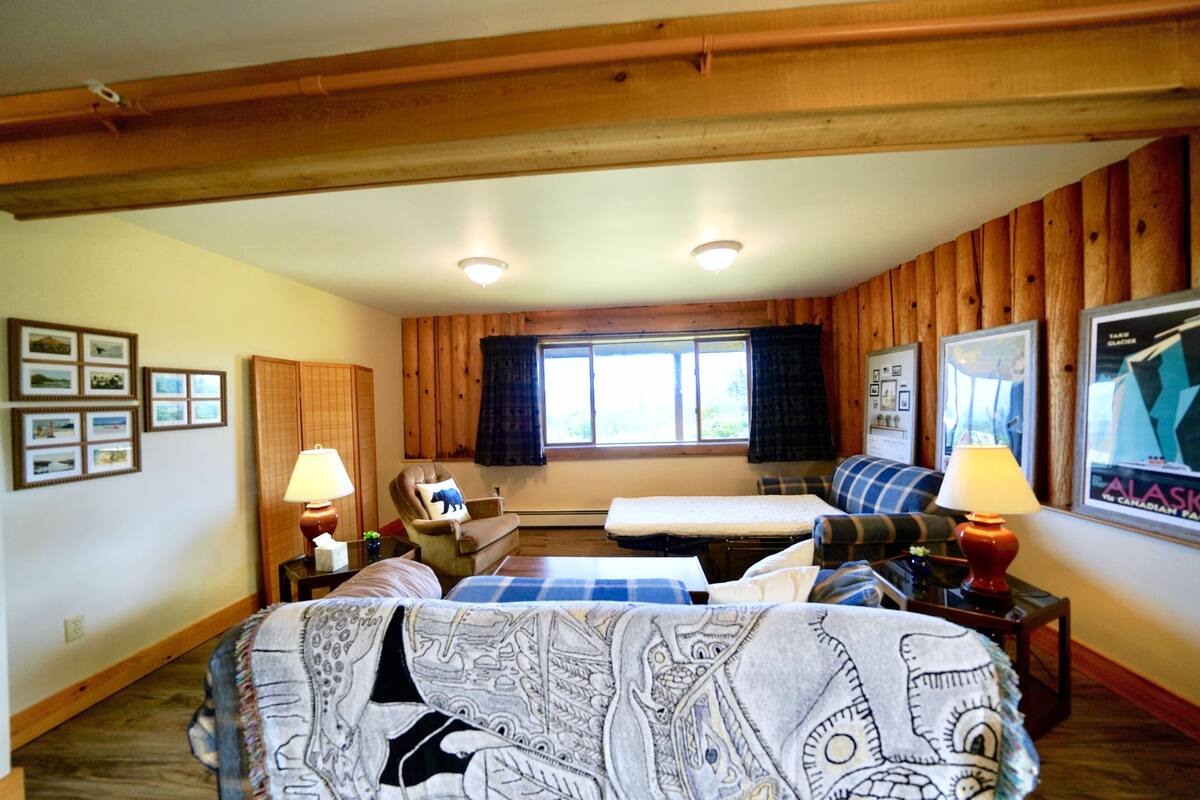 A cozy room with wooden walls, a bed with patterned bedding, a sofa, a small table with a lamp, and a window with curtains.