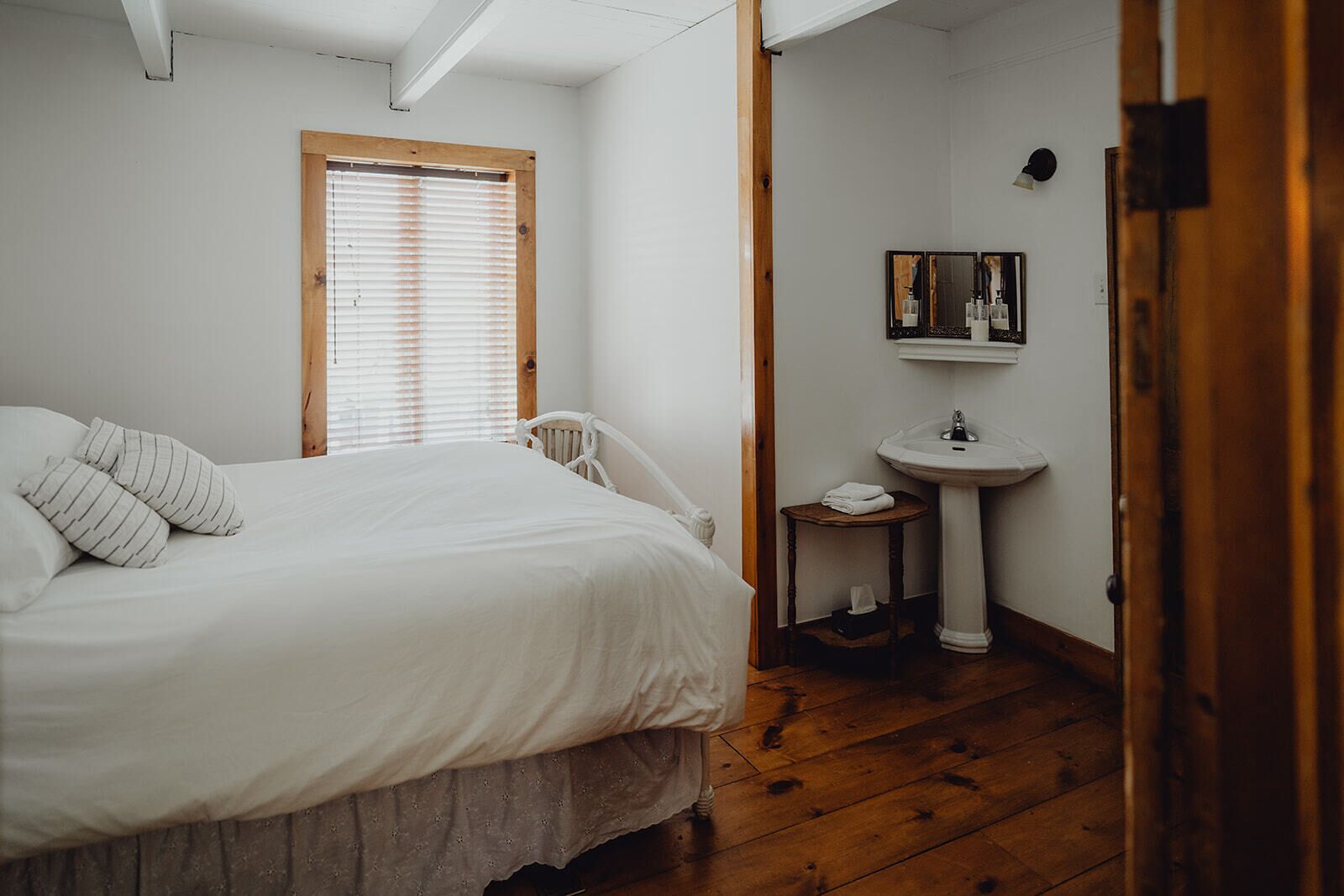 A bedroom with a bed, a wooden chair, a small bedside table with a sink, and a window with blinds.
