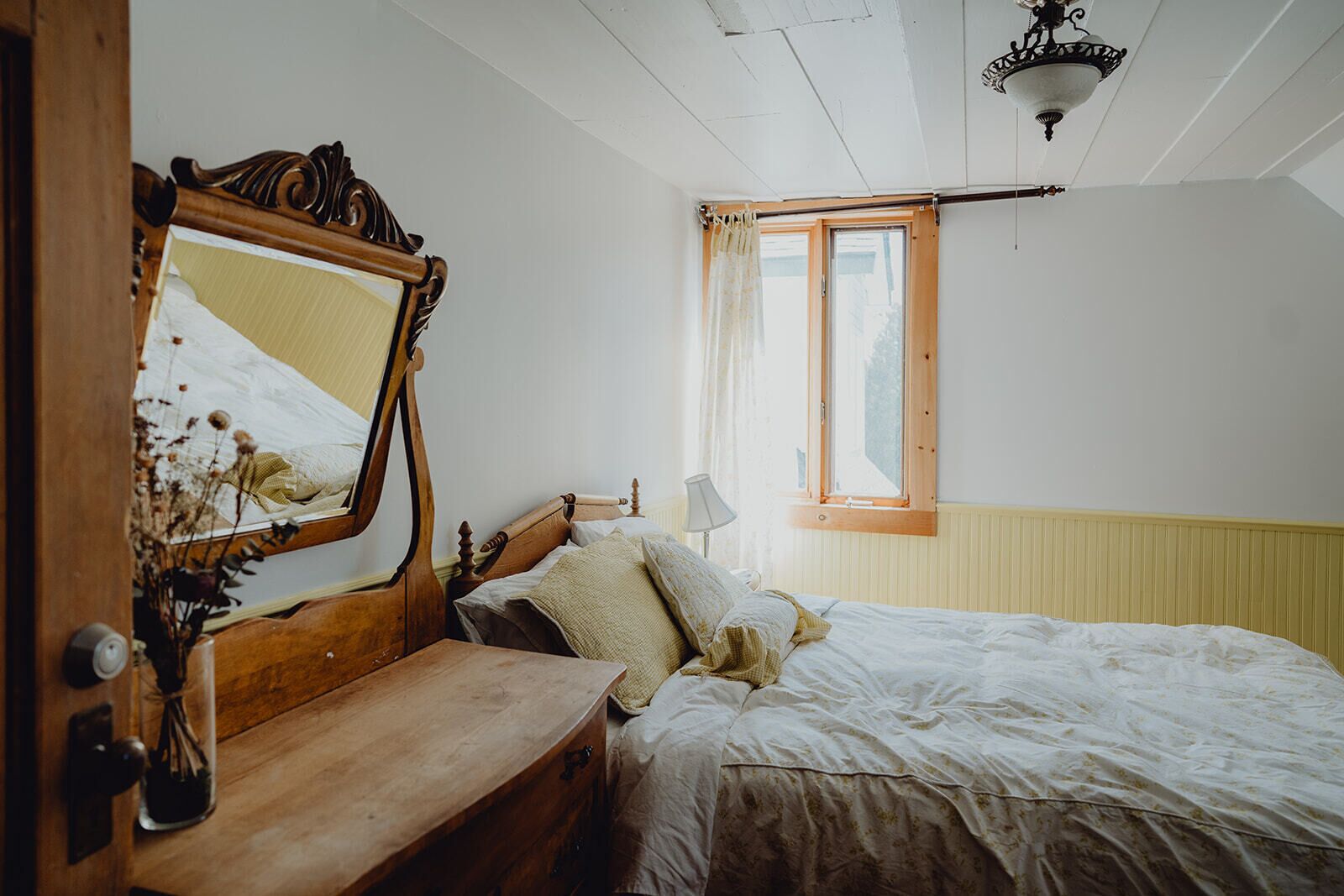 A bedroom with a wooden bed, a mirror, a bedside table with a lamp, and a window with curtains.