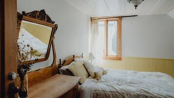 A bedroom with a wooden bed, a mirror, a bedside table with a lamp, and a window with curtains.