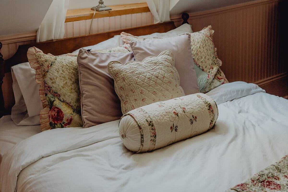 A bedroom with a sloped ceiling, a bed with multiple pillows, a bedside lamp, and a window with sheer curtains.
