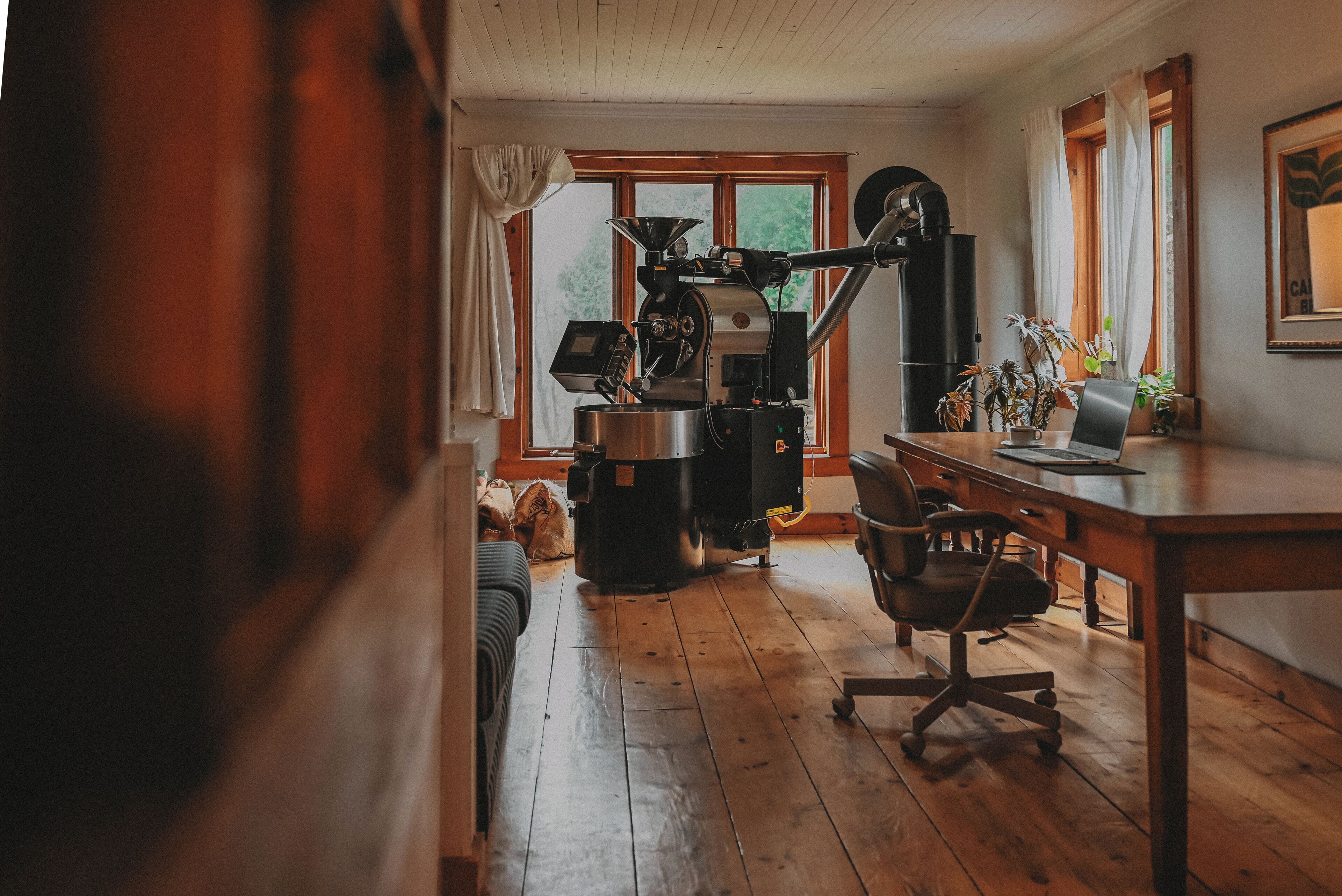 A cozy room with a wooden floor, a coffee machine, a desk with a laptop, and a window with curtains.