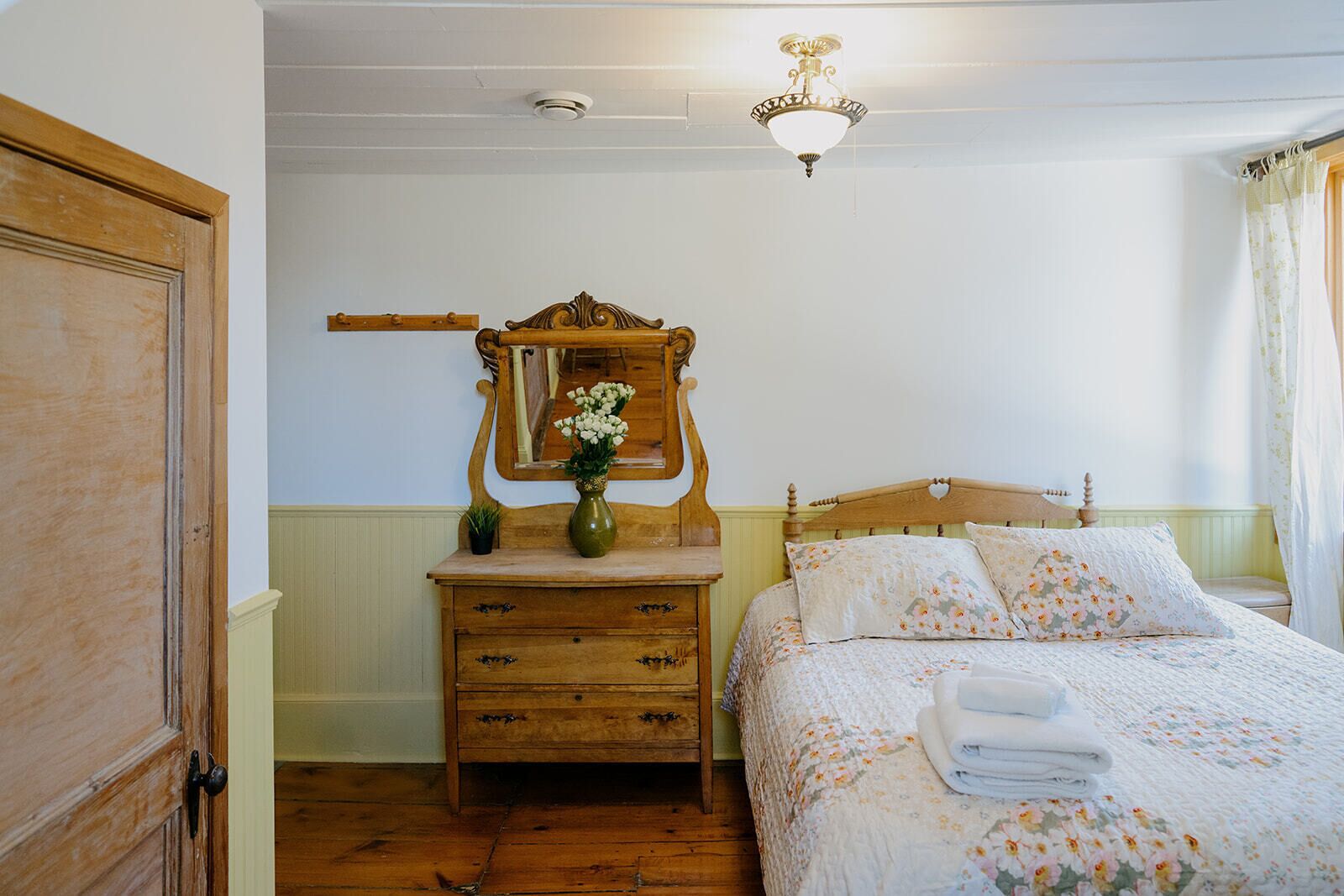 A bedroom with a wooden dresser, a mirror, a bed with floral bedding, and a ceiling light fixture.