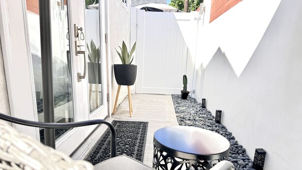 Een balkon met een rieten stoel, een klein rond tafeltje, potplanten en een witte hek.