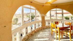 A spacious room with large arched windows, a dining area with wooden furniture, and a patterned floor.