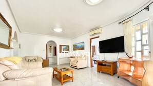 A spacious living room with a sofa, armchair, and coffee table. A flat-screen TV is mounted on the wall, and there is a window with curtains.