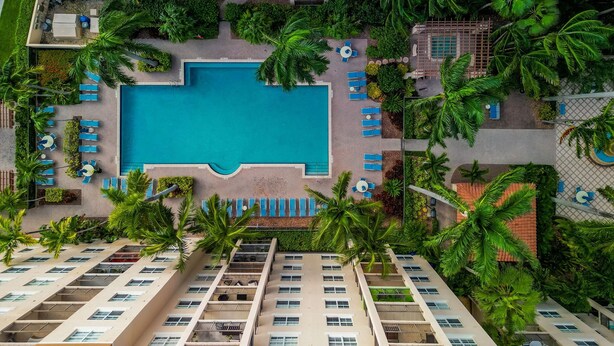 An aerial view of a hotel with a central swimming pool surrounded by lounge chairs and palm trees.