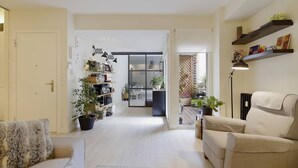 Ein Wohnzimmer mit einer Couch, Sesseln und einem Couchtisch. Es gibt Blumentöpfe, ein Bücherregal und im Hintergrund einen Essbereich.