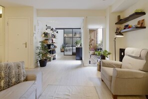 Ein Wohnzimmer mit einer Couch, Sesseln und einem Couchtisch. Es gibt Blumentöpfe, ein Bücherregal und im Hintergrund einen Essbereich.