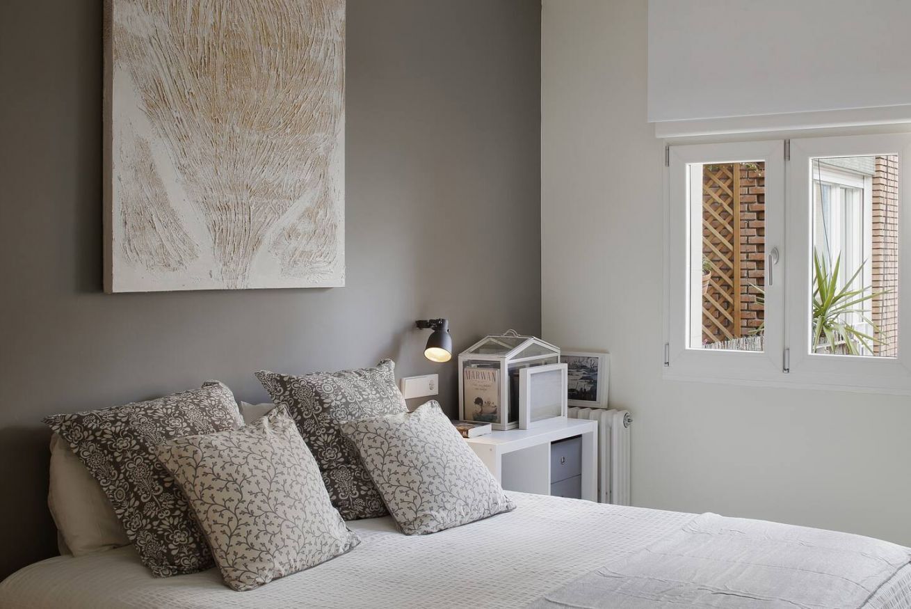 A bedroom with a bed, pillows, a wall-mounted lamp, a framed picture, and a window with a view of a brick wall and plants.