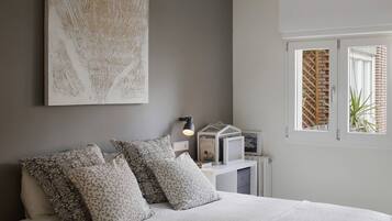 Un dormitorio con cama, almohadas, una lámpara de pared, un cuadro enmarco y una ventana que da a un muro de ladrillo y plantas.