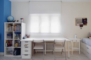 Una habitación con un escritorio blanco, dos sillas, una estantería, un radiador y una cama.