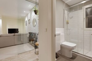 A modern bathroom with a glass-enclosed shower, a white toilet, and a glass partition separating the shower from the living area.