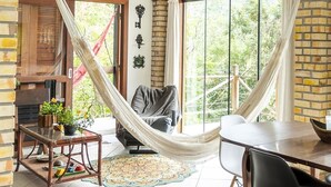 A cozy room with a hammock, a wooden table, and a chair. There is a fireplace and a large window with a view of greenery. - Local Paradisíaco NA Ilha DA Magia Loft Arcano Maior (Lagoa da Conceição Florianópolis)