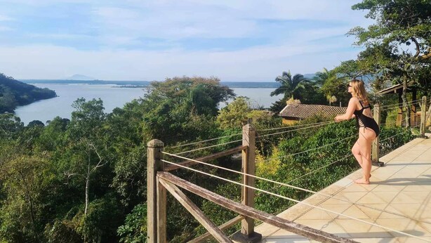 A woman standing on a wooden deck overlooking a body of water with surrounding greenery. - Local Paradisíaco NA Ilha DA Magia Loft Arcano Maior (Lagoa da Conceição Florianópolis)