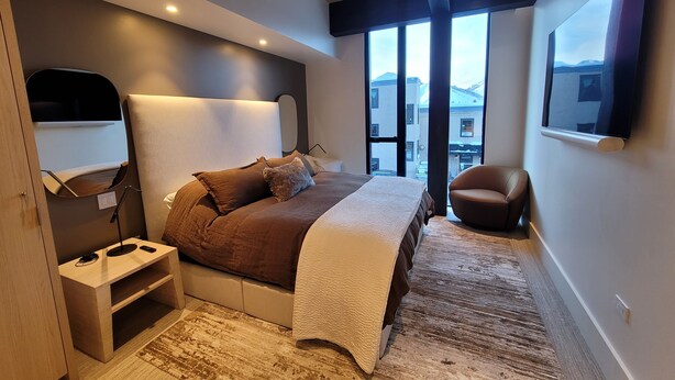 Un dormitorio moderno con una cama grande, una mesita de noche, una silla, un televisor y vistas a montañas a través de amplios ventanales.