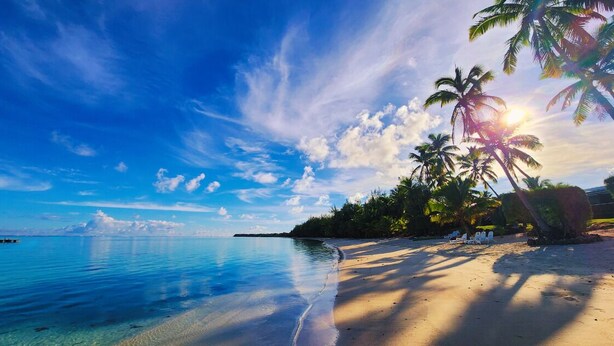 A beach with clear blue water, palm trees, and a boat in the distance. - Resort Tava'e (Aitutaki)
