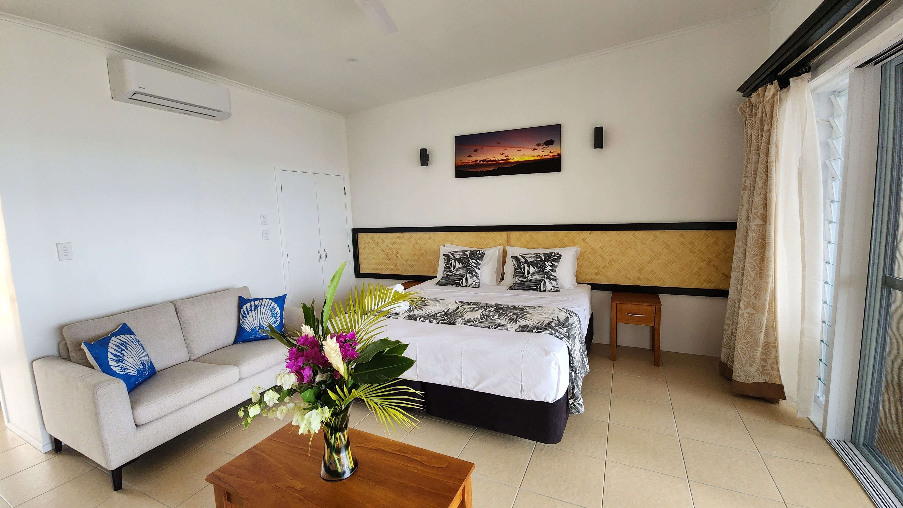 A hotel room with a bed, a sofa, a small table with a vase of flowers, and a window with curtains.