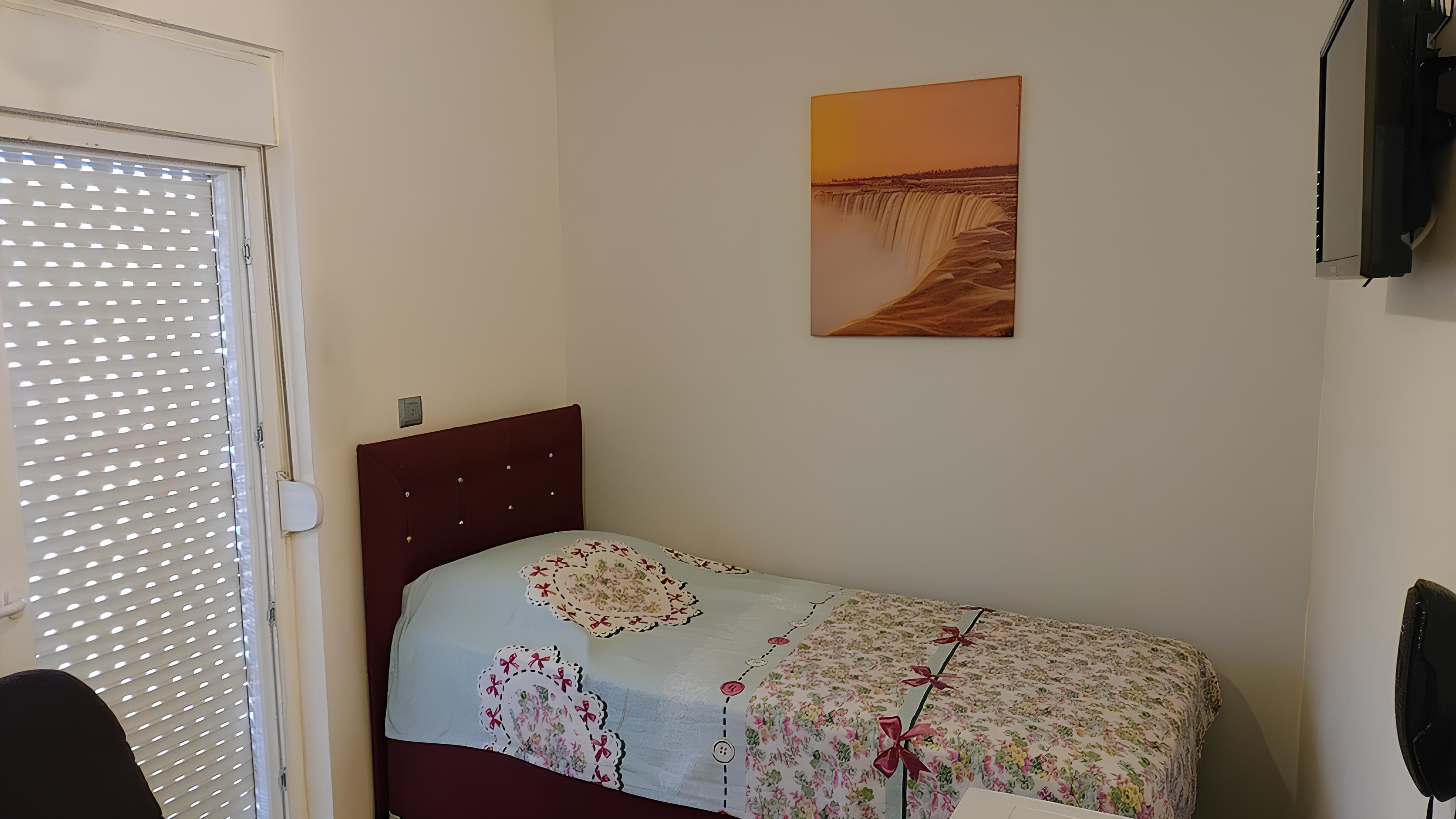 A single bed with a floral bedspread, a wall-mounted TV, and a painting of a waterfall.
