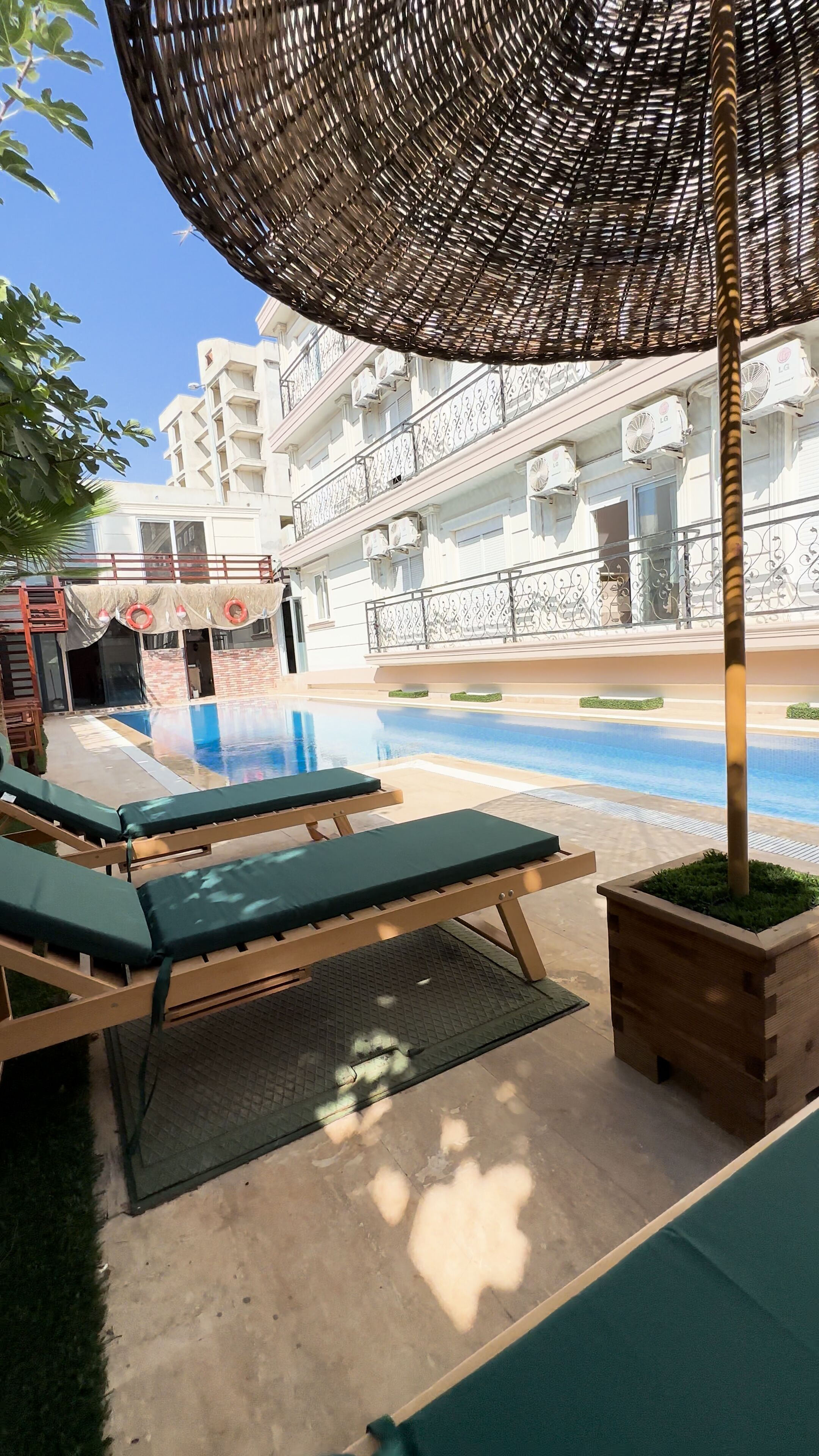 Area piscina all'aperto con lettini, un pergolato e un edificio sullo sfondo dotato di balconi.