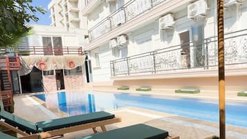 Area piscina all'aperto con lettini, un pergolato e un edificio sullo sfondo dotato di balconi.