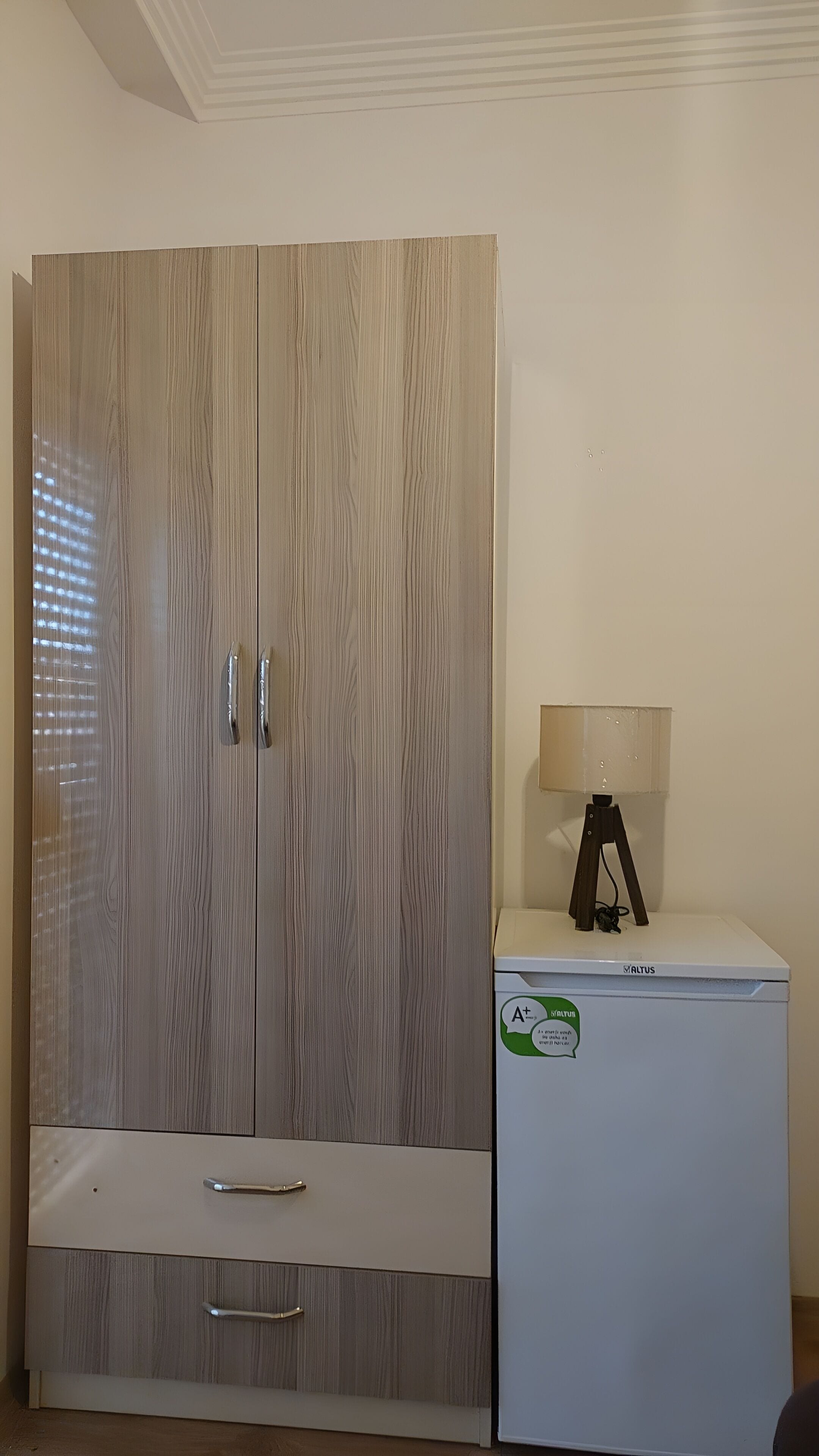 Quarto com um guarda-roupa grande, um armário branco pequeno com uma lâmpada e um teto com design decorativo.