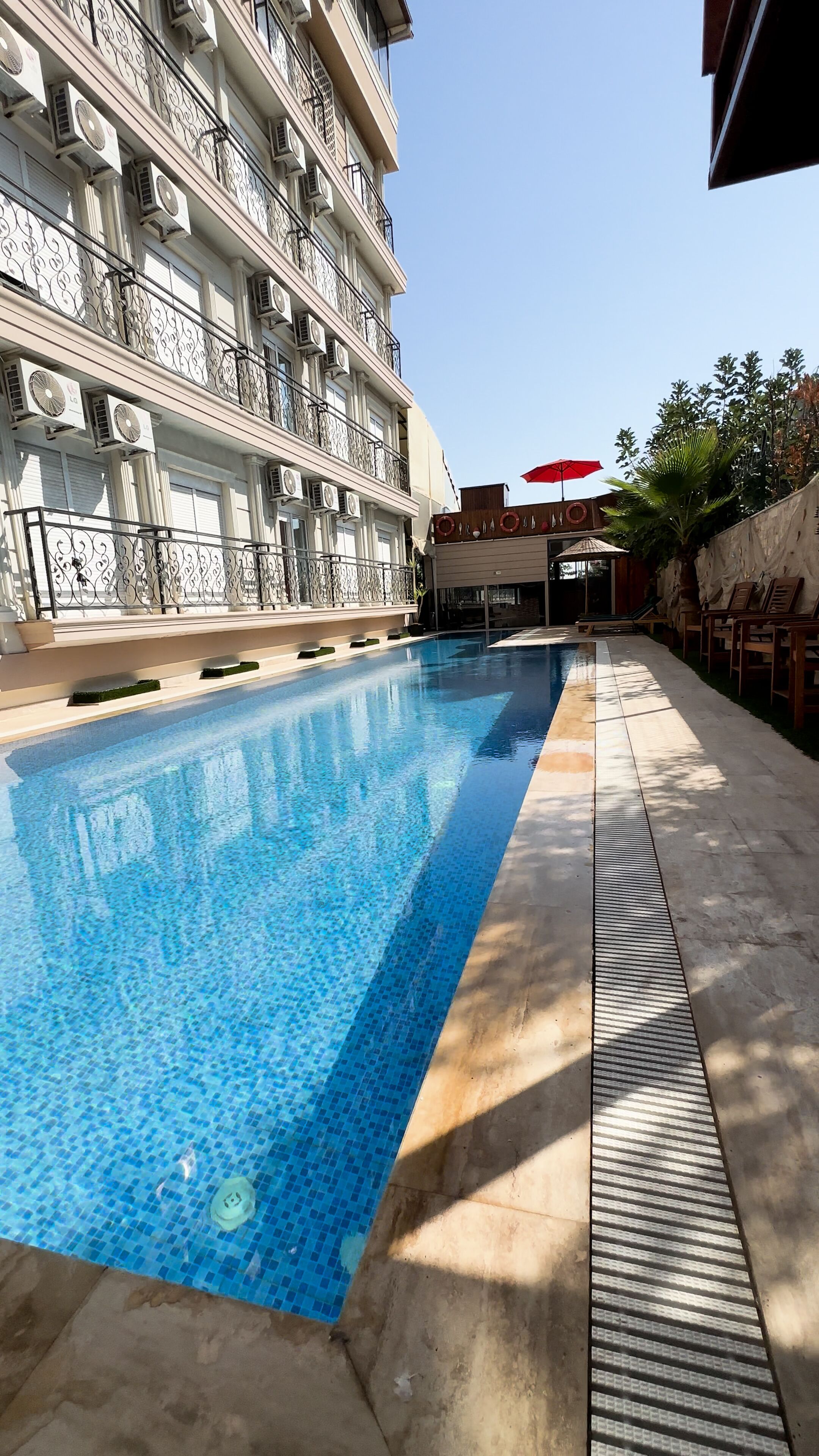 A swimming pool with a tiled edge, surrounded by a concrete deck and a building with balconies.
