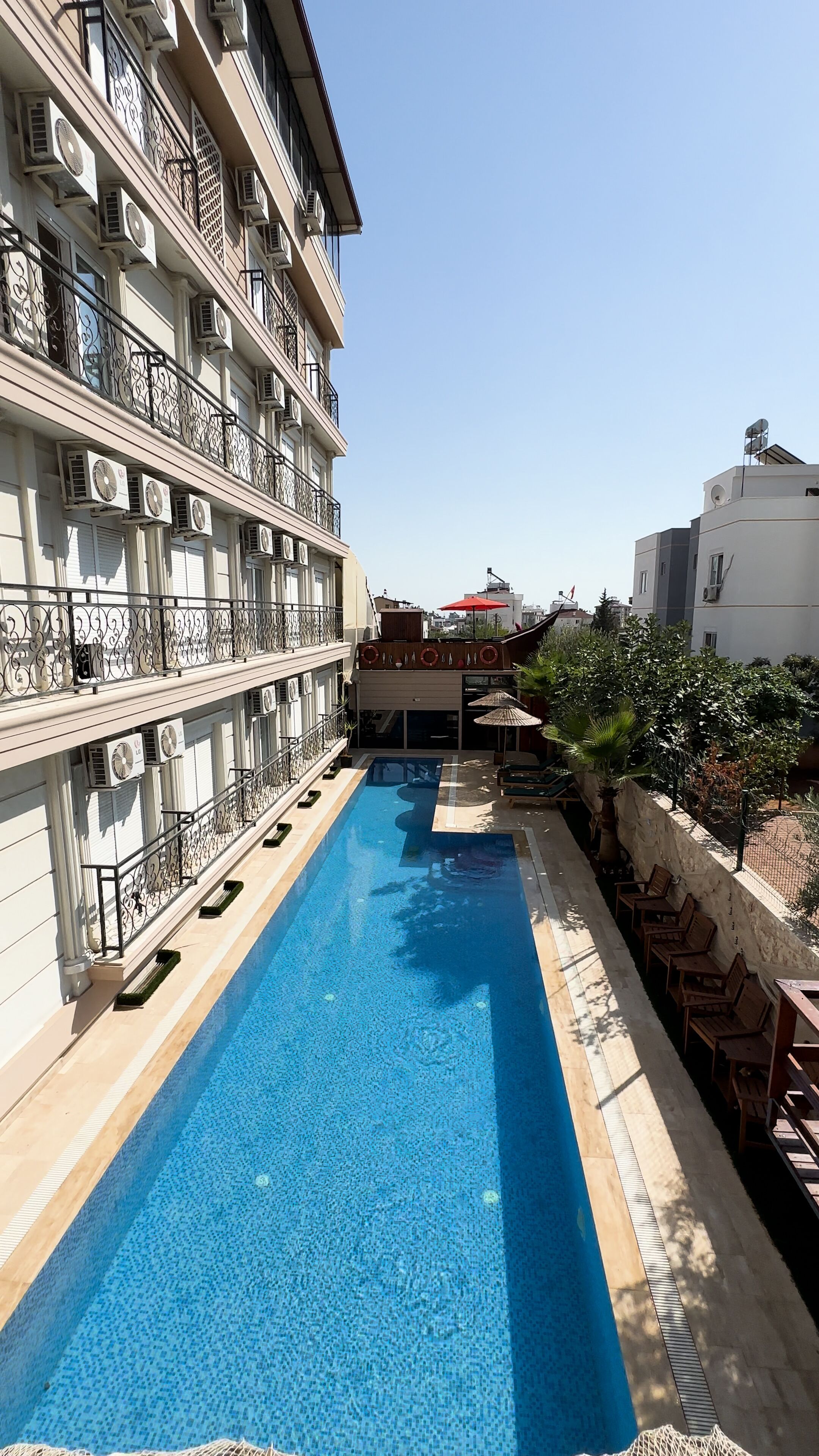 Una piscina con borde de baldosas, rodeada de sillas de descanso y un edificio con varias unidades de aire acondicionado.