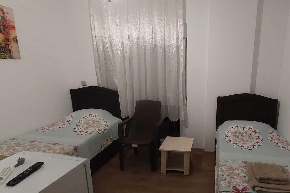 A small, simple room with two single beds, a chair, a small table, and a desk with a remote control.