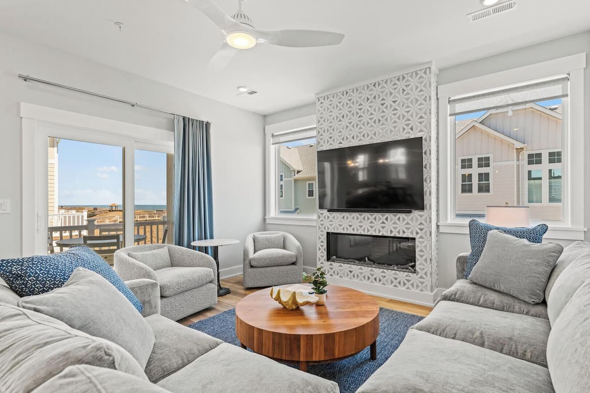 A modern living room with a fireplace, comfortable seating, and large windows offering a view of the beach.