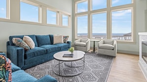 A spacious living room with a blue sofa, two armchairs, a coffee table, and large windows offering a view of the ocean. - The Tides by KEES Vacations (Kill Devil Hills)