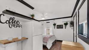 Una habitación pequeña con una cama, letrero de café, ventilador de techo y una pequeña mesa de madera.