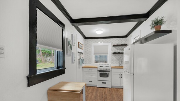 Una cocina compacta con armarios blancos, una mesa de madera y un ventanal con persianas.