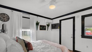 Un dormitorio con cama, cortinas blancas, ventilador de techo, televisor montado y una puerta.