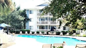 Ein Pool mit Liegestühlen und Sonnenschirmen, umgeben von Grünflächen und einem mehrstöckigen Gebäude.