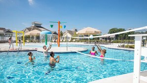 Ein Schwimmbecken mit Volleyballnetz, Wasserrutschen und einem überdachten Sitzbereich.