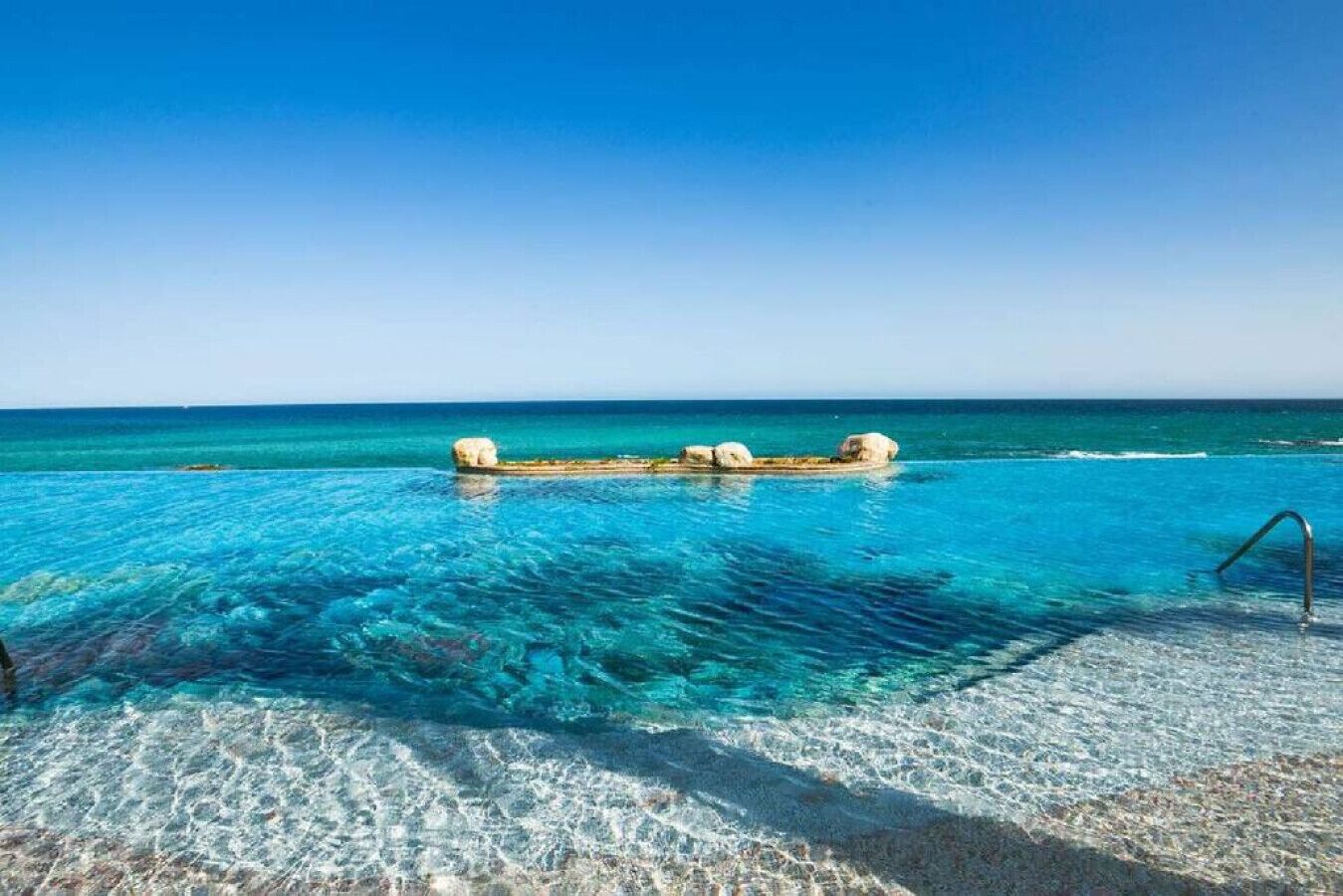 Une piscine d’un bleu éclatant, bordée d’une margelle en pierre et dotée d’un escalier en métal, entourée d’une plage de sable et de l’océan.